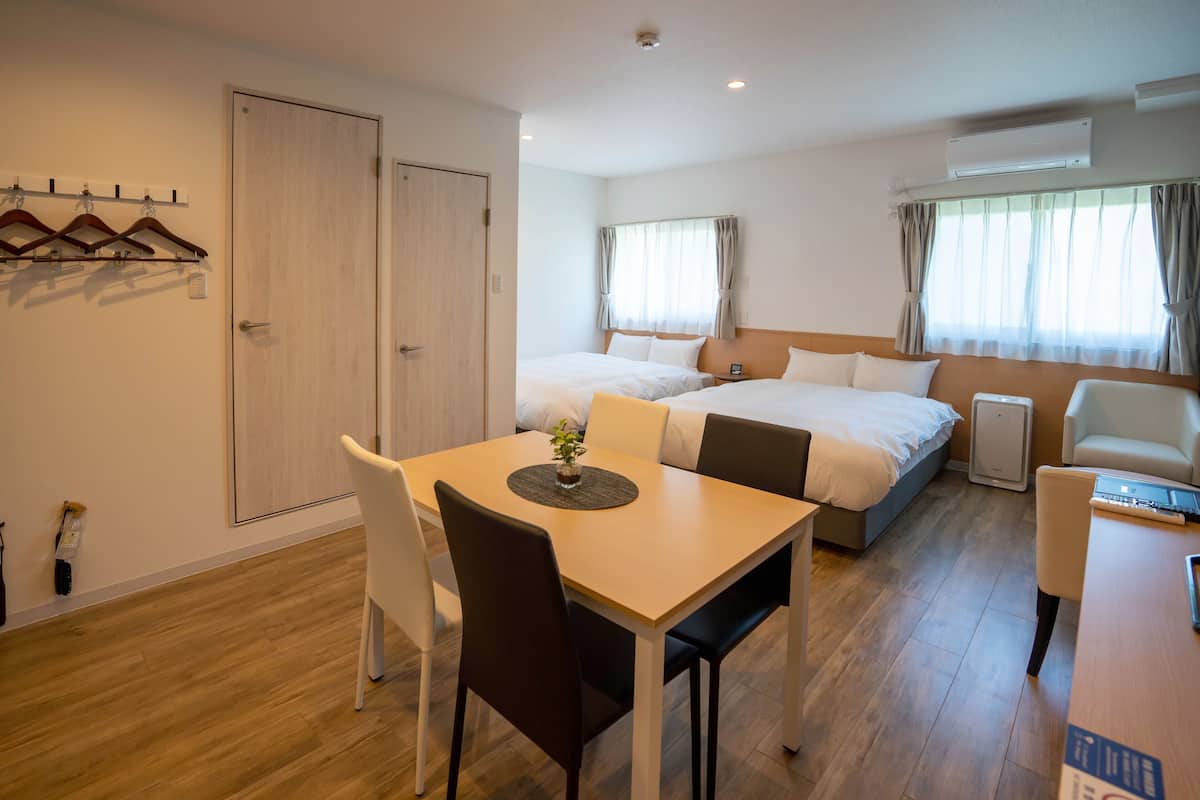 A hotel room with a wooden floor, a dining table with chairs, a bed with a headboard, a bedside table, and a window with curtains.