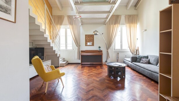 Uma sala de estar espaçosa com piso em madeira em padrão herringbone, uma poltrona amarela, um sofá cinza, um piano e uma escada com vista para o andar superior.
