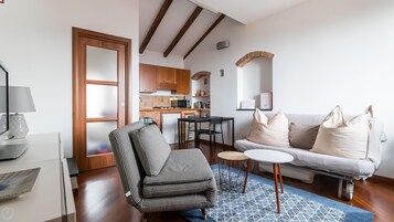 A compact living space with a kitchenette, a sofa with cushions, a patterned armchair, a small dining table, and a lamp.