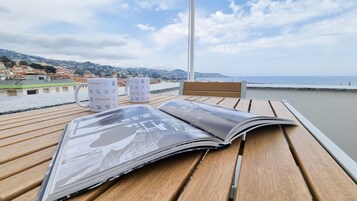 A wooden table with a magazine and two mugs on a balcony overlooking a coastal town.