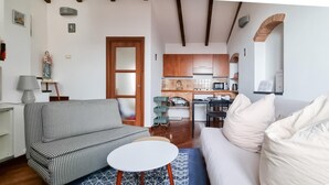 A living room with a sofa, a white armchair, and a small table. There is a kitchen in the background with wooden cabinets and a microwave. - Italianway - Costiglioli 8 B (Sanremo)