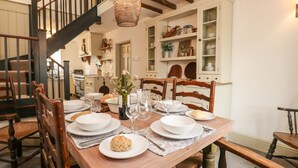Un comedor con una mesa de madera dispuesta para cuatro personas, rodeada de sillas de madera. Se ve una escalera con barandilla oscura al fondo.