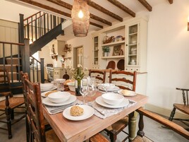 Un comedor con una mesa de madera dispuesta para cuatro personas, rodeada de sillas de madera. Se ve una escalera con barandilla oscura al fondo.