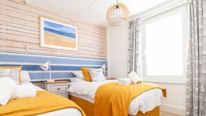 A hotel room with two beds, a wooden headboard, a beach-themed wall art, a window with curtains, and a hanging light fixture.