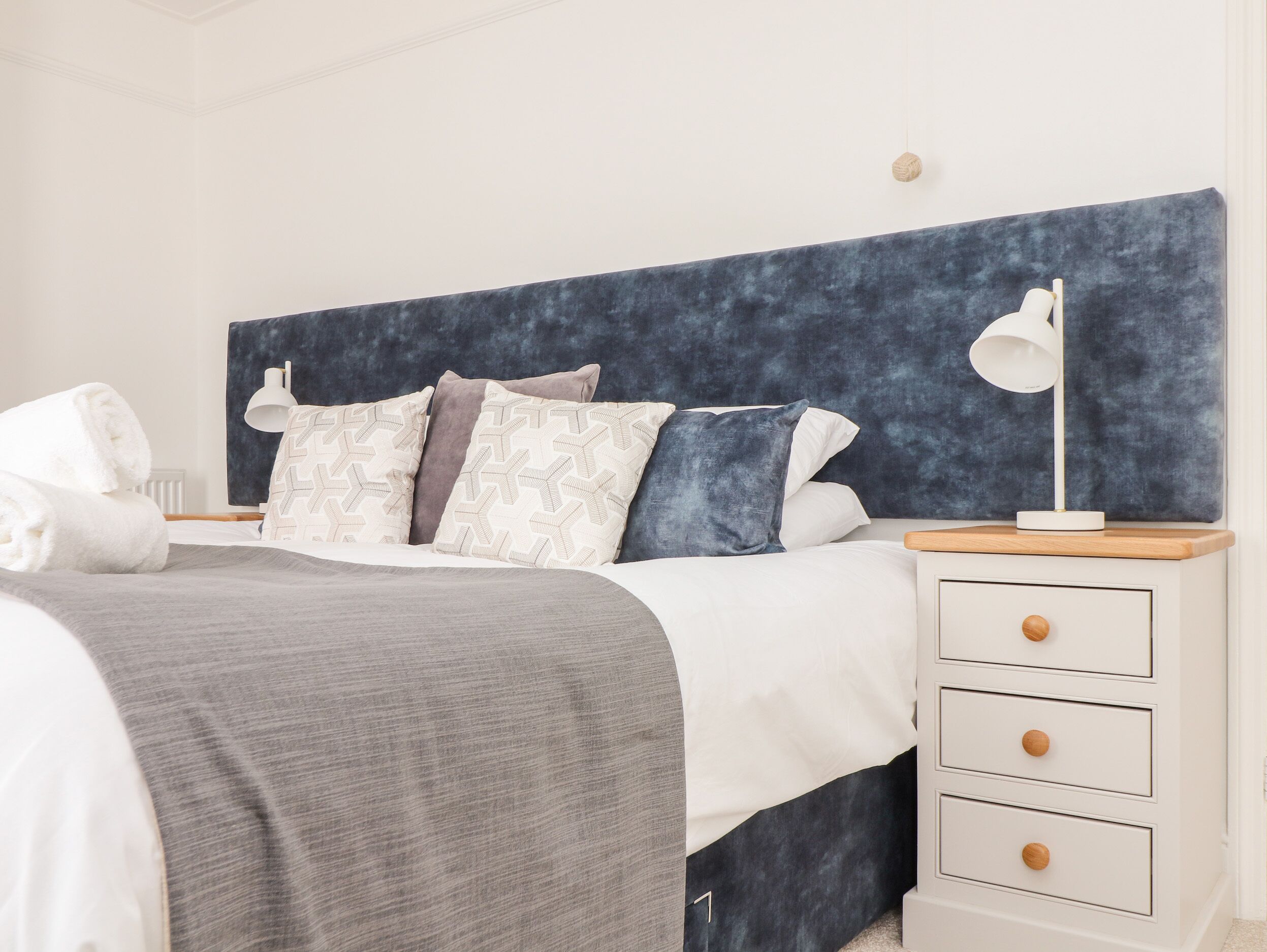 A neatly made bed with a grey blanket, a white pillow, and a blue headboard. There is a bedside table with a lamp and a drawer.