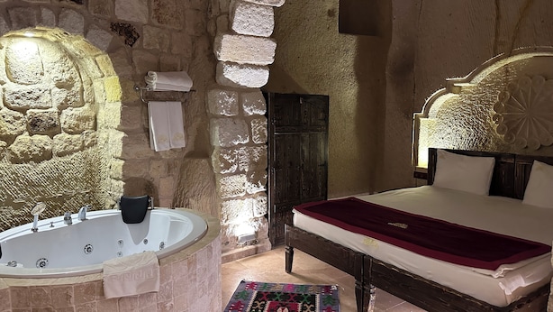 Un dormitorio con paredes de piedra, una bañera grande, una cama de madera con colcha roja y una alfombra estampada.