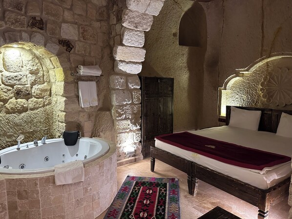 A stone-walled bedroom with a large bathtub, a wooden bed with a red bedspread, and a patterned rug. - Under Cave Hotel (Ürgüp)