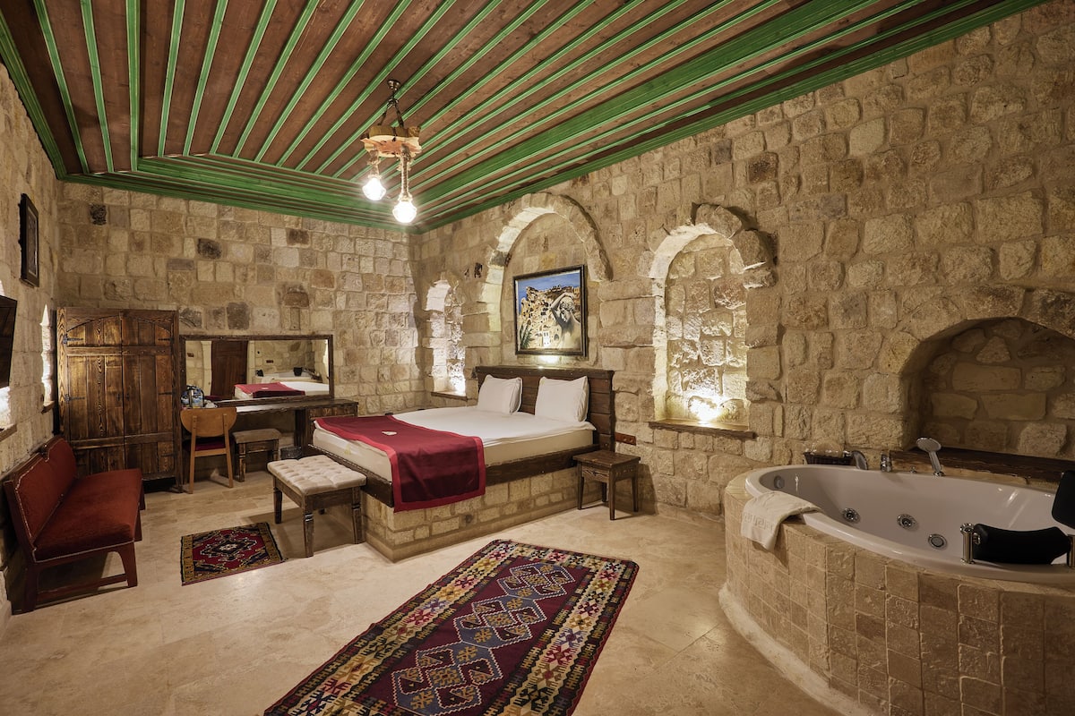 A stone-walled bedroom with a large bed, a wooden desk, a red bench, a patterned rug, and a bathtub.