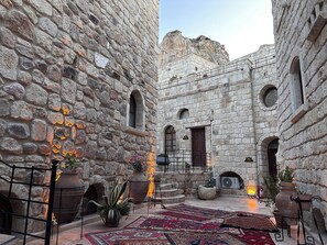 A courtyard with stone walls, a fire extinguisher, a large potted plant, and a patterned rug. - Under Cave Hotel (Ürgüp)