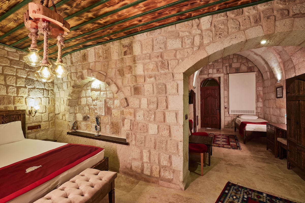 A room with a stone wall, a bed with a red bedspread, a wooden bench, and a hanging light fixture.