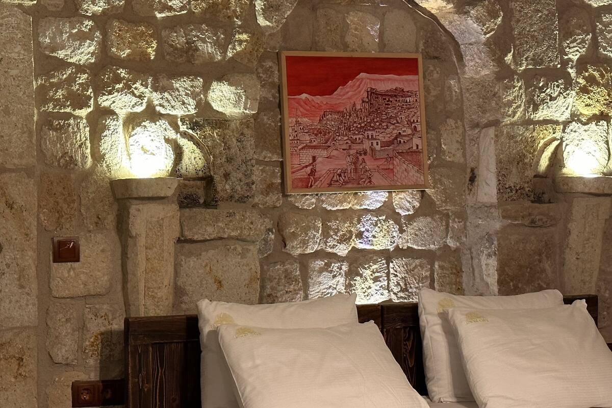 A room with stone walls, a bed with white linens, and a framed picture of a town on the wall.