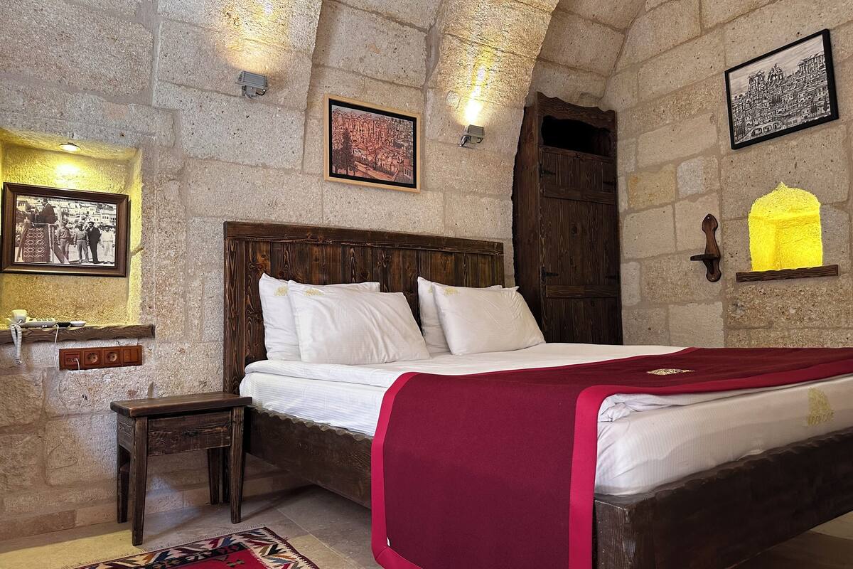 A stone-walled bedroom with a wooden bed, a nightstand, a red throw blanket, and two framed pictures.
