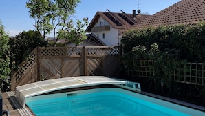 Una piscina con cubierta de madera, cerca hay una valla de madera y, al fondo, una casa con techo de tejas.