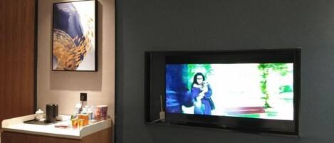A television mounted on a dark wall, a wooden panel to the left, and a small counter with various items.
