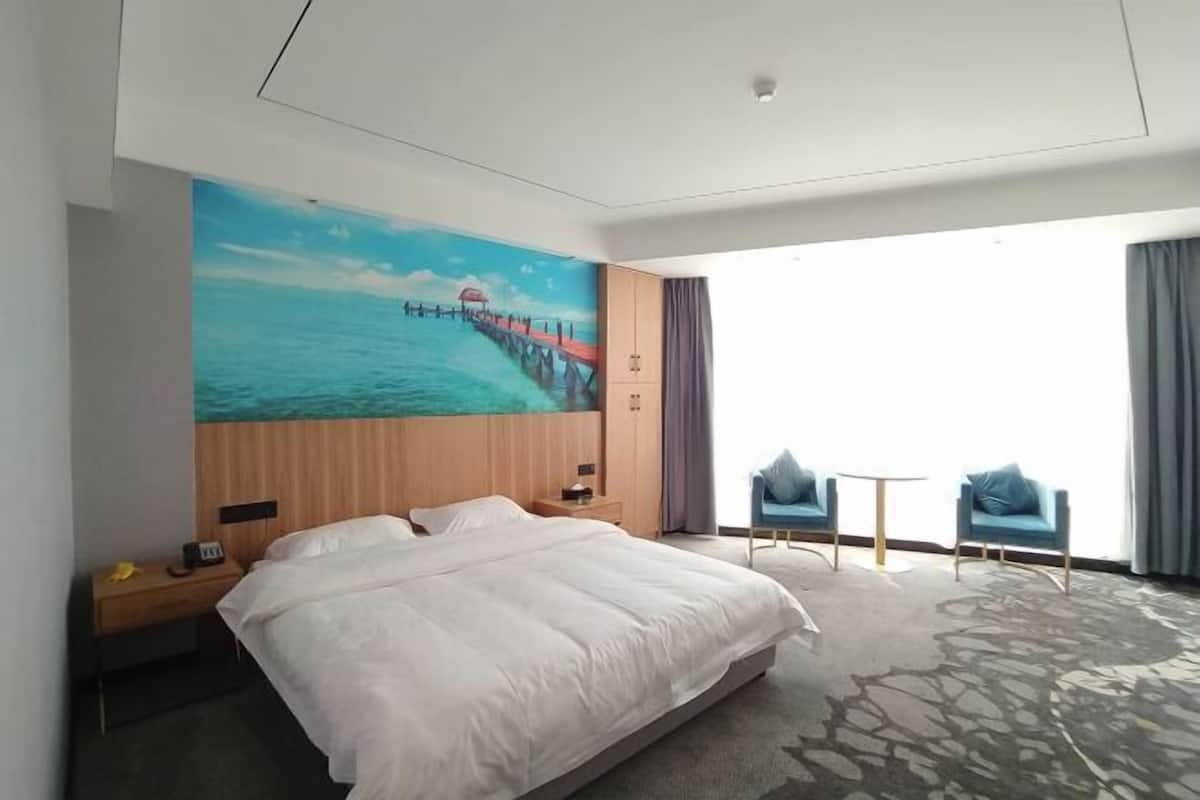 A hotel room with a large bed, a nightstand, a desk, two chairs, a small table, and a mural of a pier extending into a blue sea.