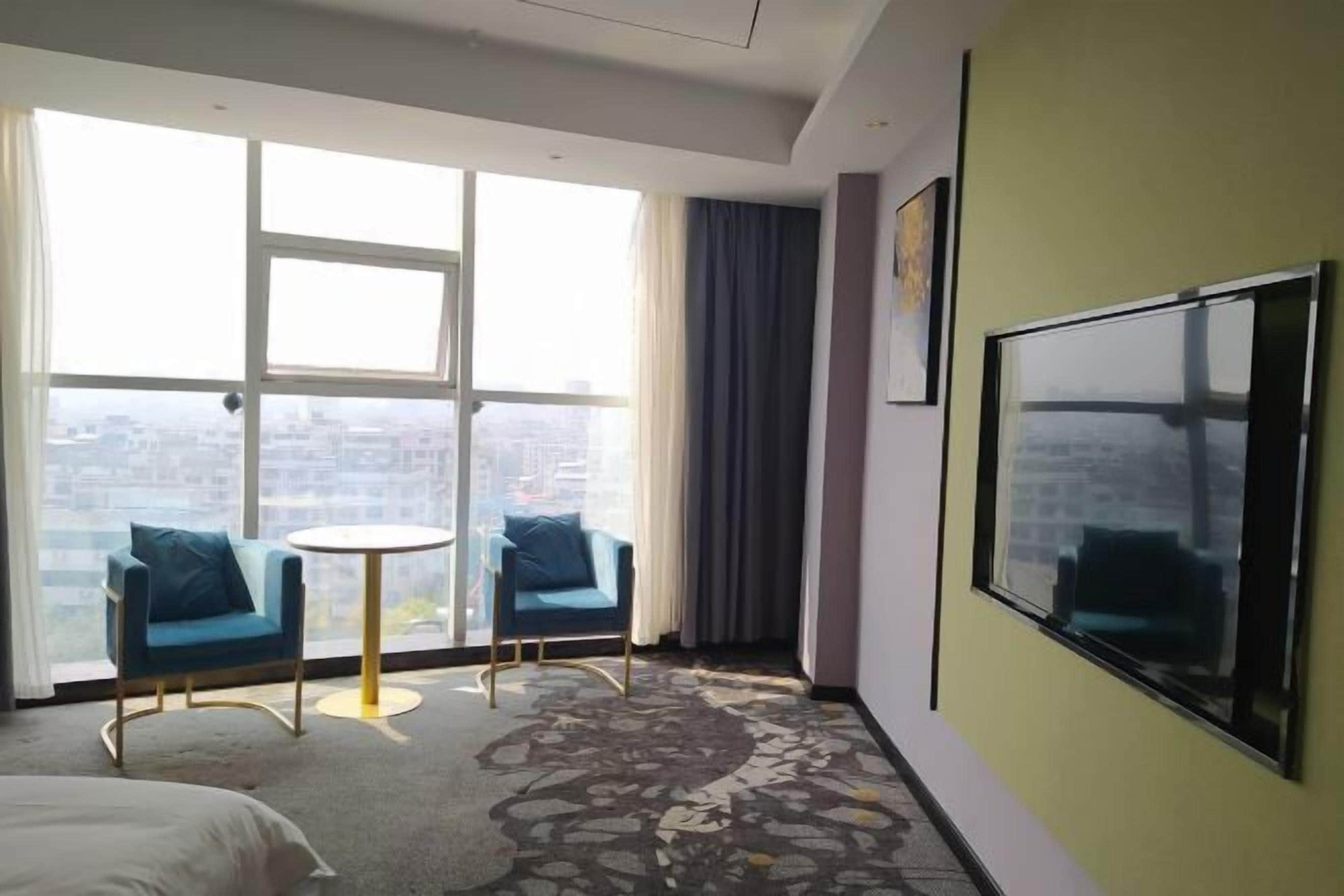 Habitación de hotel con un ventanal grande, dos sillones azules, una pequeña mesa redonda y un televisor de pantalla plana grande fijado a la pared.