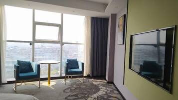 A hotel room with a large window, two blue armchairs, a small round table, and a large flat-screen TV mounted on the wall.