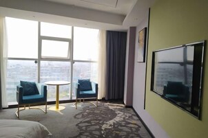 Habitación de hotel con un ventanal grande, dos sillones azules, una pequeña mesa redonda y un televisor de pantalla plana grande fijado a la pared.