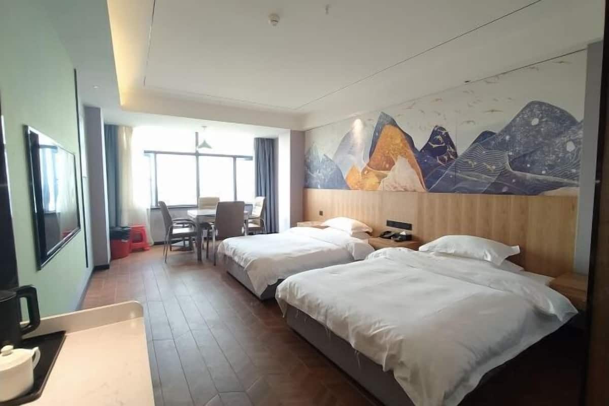 A hotel room with two beds, a desk, and a chair. There is a large window providing natural light.