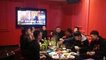 A group of people seated around a table with food and drinks, a television screen in the background, and red walls.