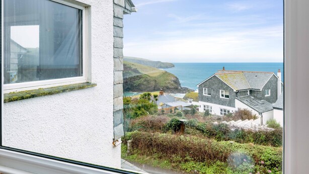 Une vue sur la côte, depuis une fenêtre donnant sur un bâtiment blanc aux fondations en pierre, une zone herbeuse avec des arbustes et une vue sur la mer avec des falaises.
