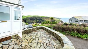 Une entrée pavée de pierres, avec une porte blanche, un mur de pierre et une vue sur un paysage côtier avec des collines et une maison au loin.