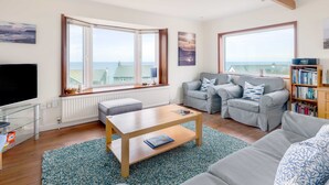 Un salon spacieux doté d’une grande fenêtre offrant une vue sur la mer, d’un téléviseur à écran plat, d’une table basse en bois et d’une bibliothèque.