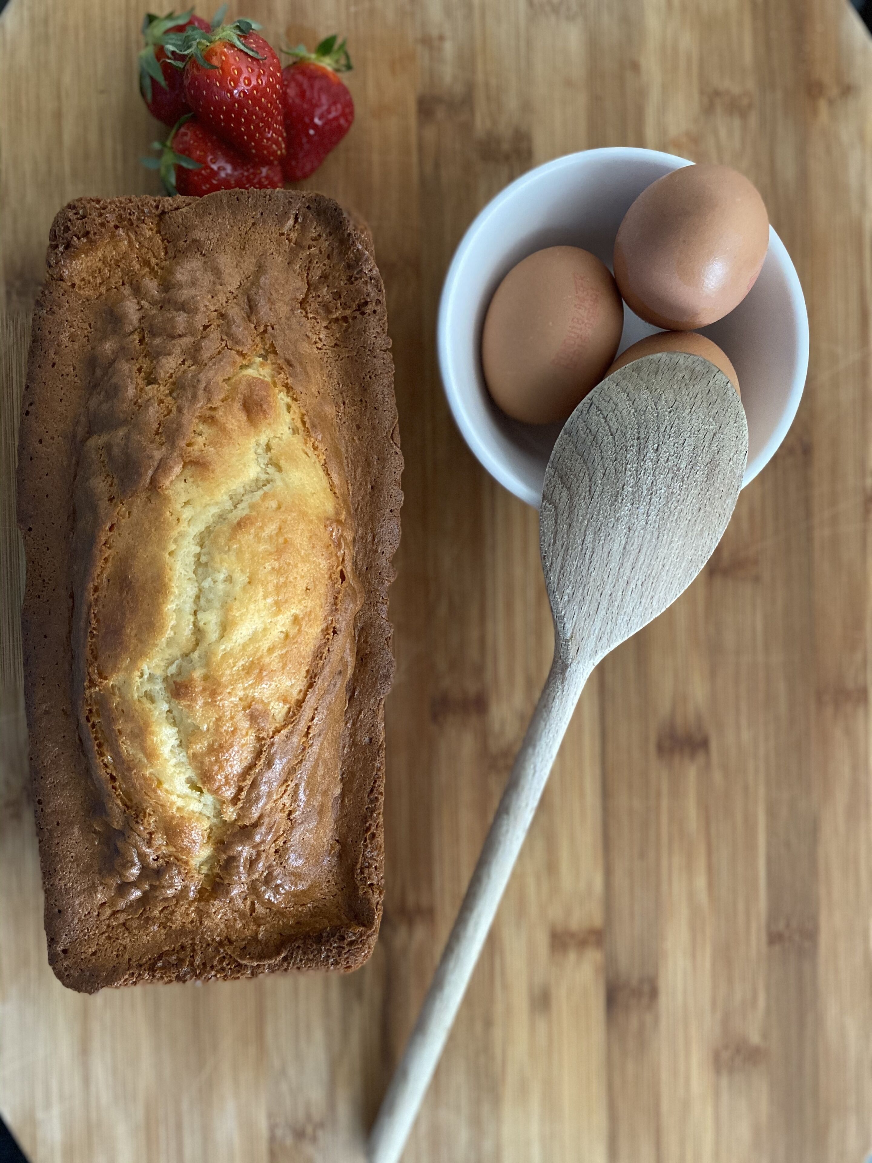 A loaf of bread on a wooden surface with a bowl of eggs and a spoon nearby.
