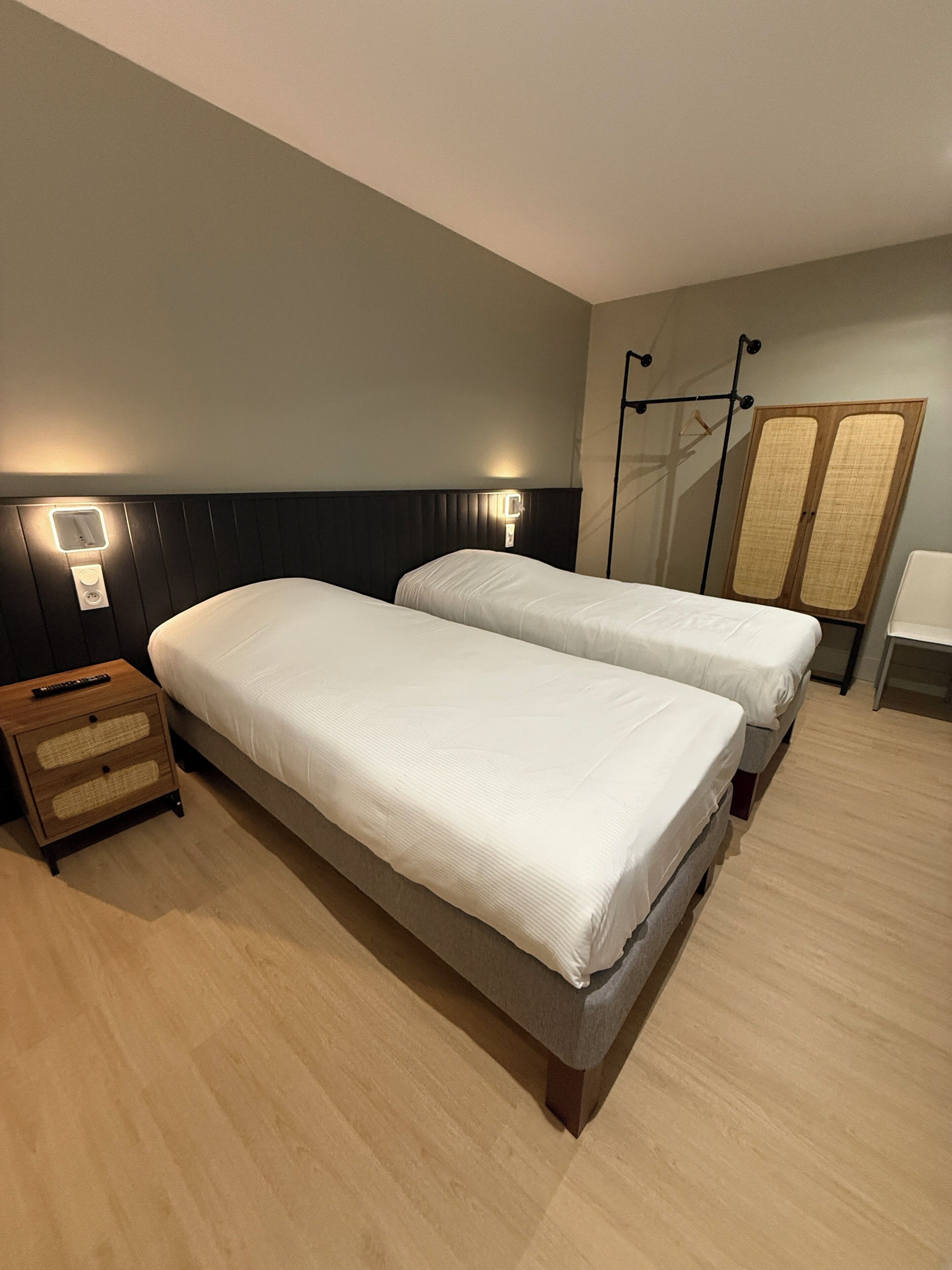 A hotel room with two single beds, a wooden nightstand, a wall-mounted light, and a wardrobe.
