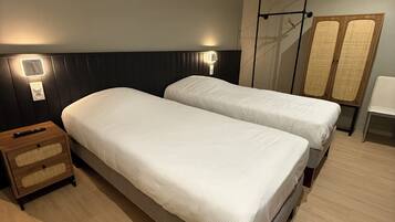 A hotel room with two single beds, a wooden nightstand, a wall-mounted light, and a wardrobe.