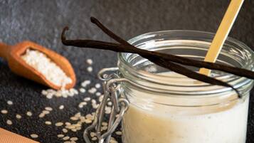 A glass jar with a metal clasp and a wooden spoon, containing a white liquid, possibly milk, with vanilla and chocolate sticks.