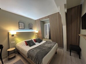 A modern bedroom with a bed, bedside tables, a small chair, and a wall-mounted shelf. - Hotel SPA Piscine le Petit Castel (Beuzeville)