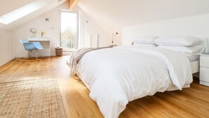 A bedroom with a sloped ceiling, a bed with white bedding, a desk with a chair, and a window providing natural light. - Apple Blossom Barn (Penzance)