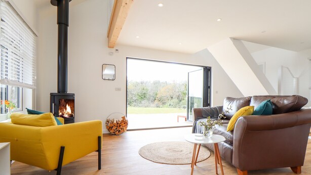 A modern living room with a wood-burning stove, a sofa, a coffee table, and a view through a glass door to the outdoors. - Apple Blossom Barn (Penzance)