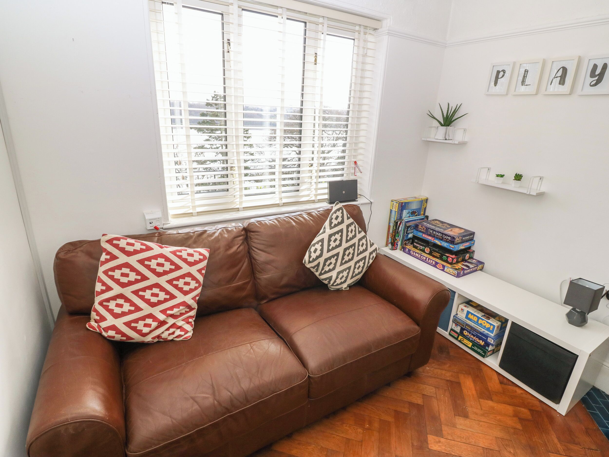 A brown leather sofa with patterned cushions, a white wall with framed artwork, a small shelf with plants and books, and a wooden herringbone floor.