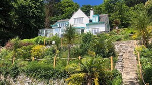 Une maison avec un escalier en pierre menant à l’entrée, entourée d’une végétation luxuriante et d’une clôture en corde.