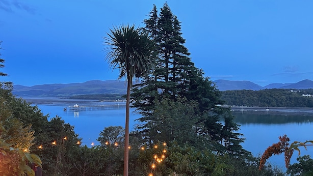 Une terrasse agrémentée d’un foyer, entourée de verdure et offrant une vue sur un lac, avec des montagnes en toile de fond.