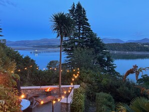 A patio with a fire feature, surrounded by greenery and overlooking a lake with mountains in the background. - Afallon (Menai Bridge)