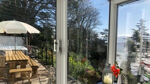 Une porte vitrée donne accès à un espace patio aménagé avec un jacuzzi, du mobilier en bois et une vue sur un paysage montagneux.