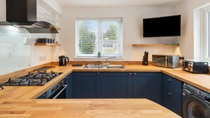 A modern kitchen with a built-in stove, sink, and dishwasher, a window with blinds, and a flat-screen TV mounted on the wall. - Falcon House in Pembroke Dock (Pembroke Dock)
