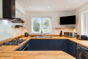 A modern kitchen with a gas stove, sink, and built-in dishwasher. There is a window with blinds, a flat-screen TV mounted above the counter, and a wooden countertop. - Falcon House in Pembroke Dock (Pembroke Dock)