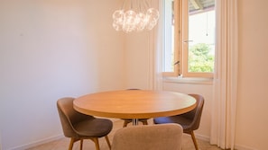 A round wooden dining table with four chairs, a hanging light fixture, and a window with curtains.