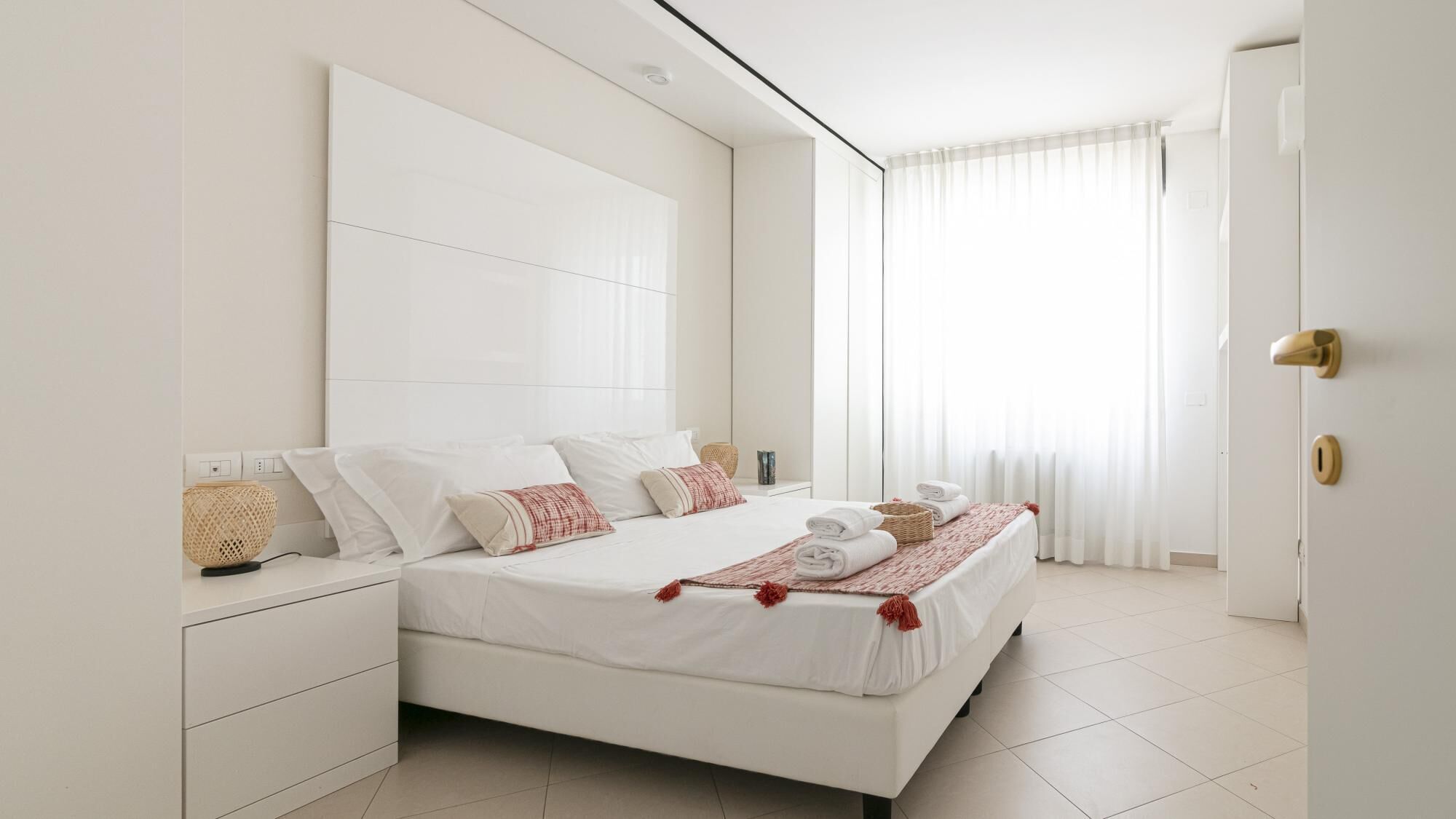 A modern bedroom with a large bed, white walls, and a large window with sheer curtains.