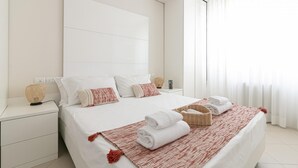 Una cama bien hecha con ropa de cama blanca y detalles rojos, una mesita de noche con lámpara y una ventana con cortinas translúcidas.