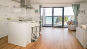 Une cuisine moderne dotée d’un îlot central, un coin repas offrant une vue sur la mer, et un espace de vie avec un canapé et un fauteuil.