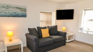 A living room with a grey sofa, two bedside tables with lamps, a flat-screen TV, and a window with blinds.