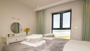 A bedroom with a bed, a dresser, a mirror, a vase with flowers, and a view of the beach through a window. - Fistral Watch (Newquay)
