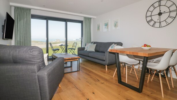 A modern living room with a sofa, a dining table with chairs, and a large window with a view of the beach. - Fistral Watch (Newquay)