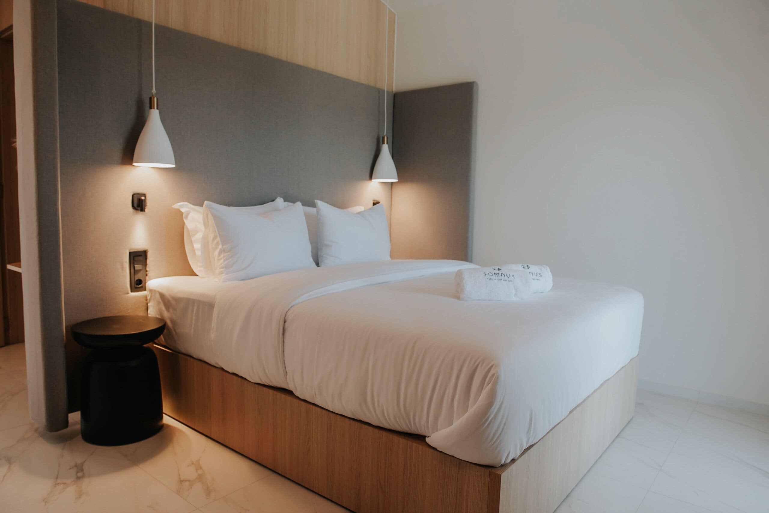 Une chambre moderne avec une tête de lit en bois, un lit simple avec une literie blanche, deux lampes suspendues et un petit tabouret noir.