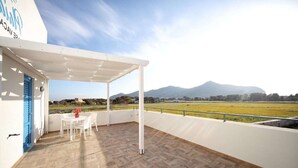 A covered outdoor area with a table and chairs, a white door, and a view of a field and mountains. - Holiday Home in Favignana near Beach (Favignana)